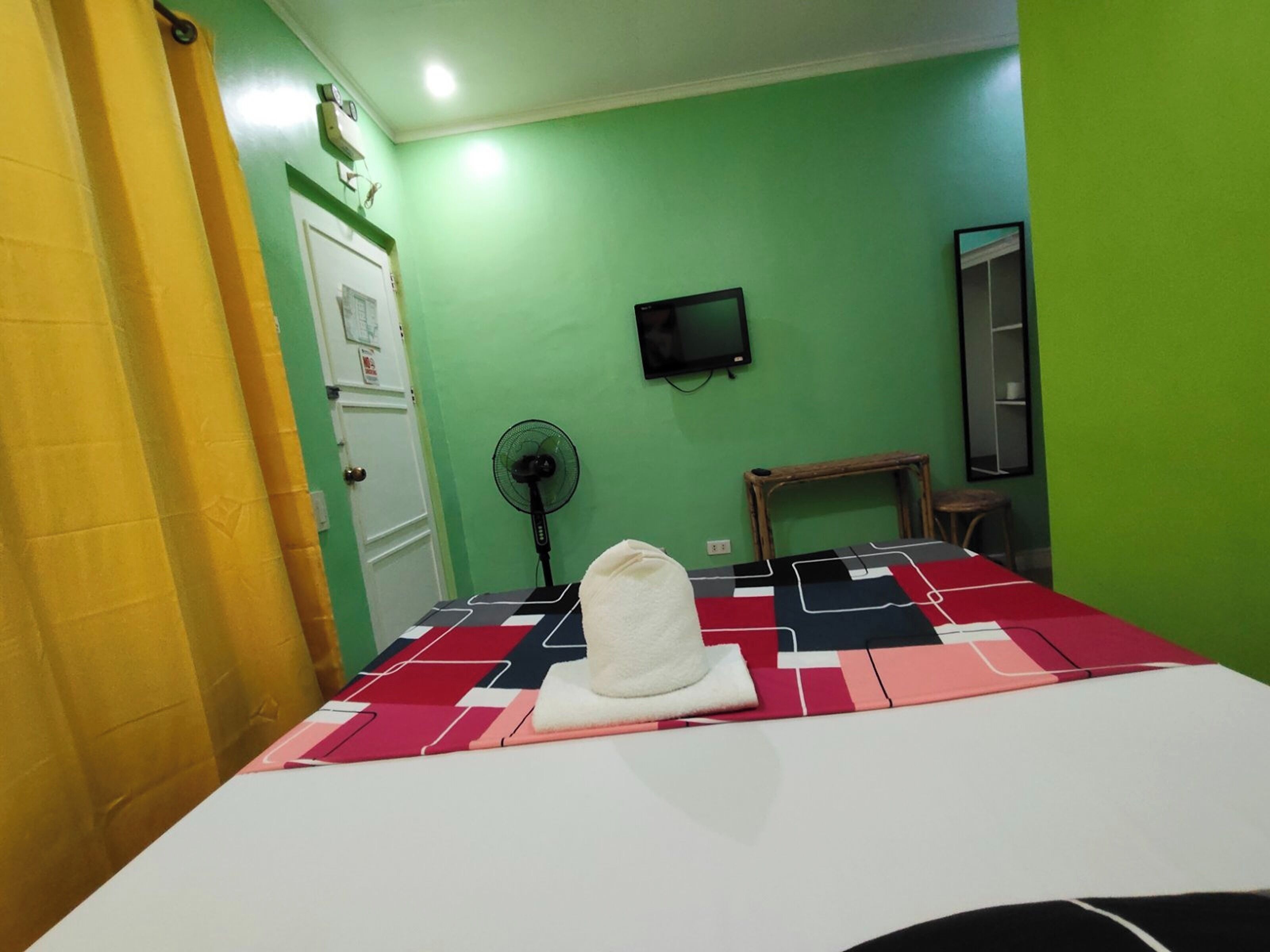 A room with a bed, a fan, a television on the wall, and a door.