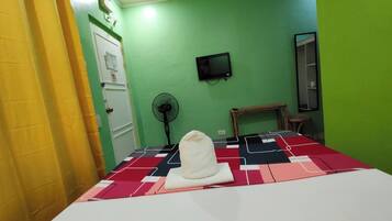 A room with a bed, a fan, a television on the wall, and a door.
