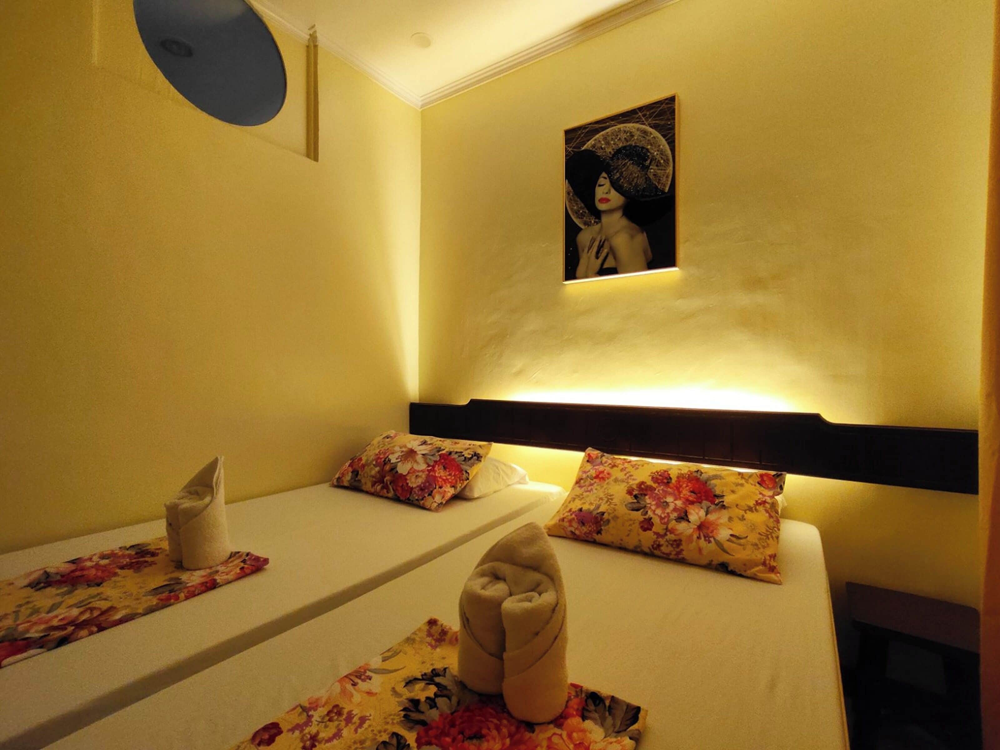Habitación de hotel con dos camas, cada una con almohadas estampadas con motivos florales y una toalla doblada con cuidado.