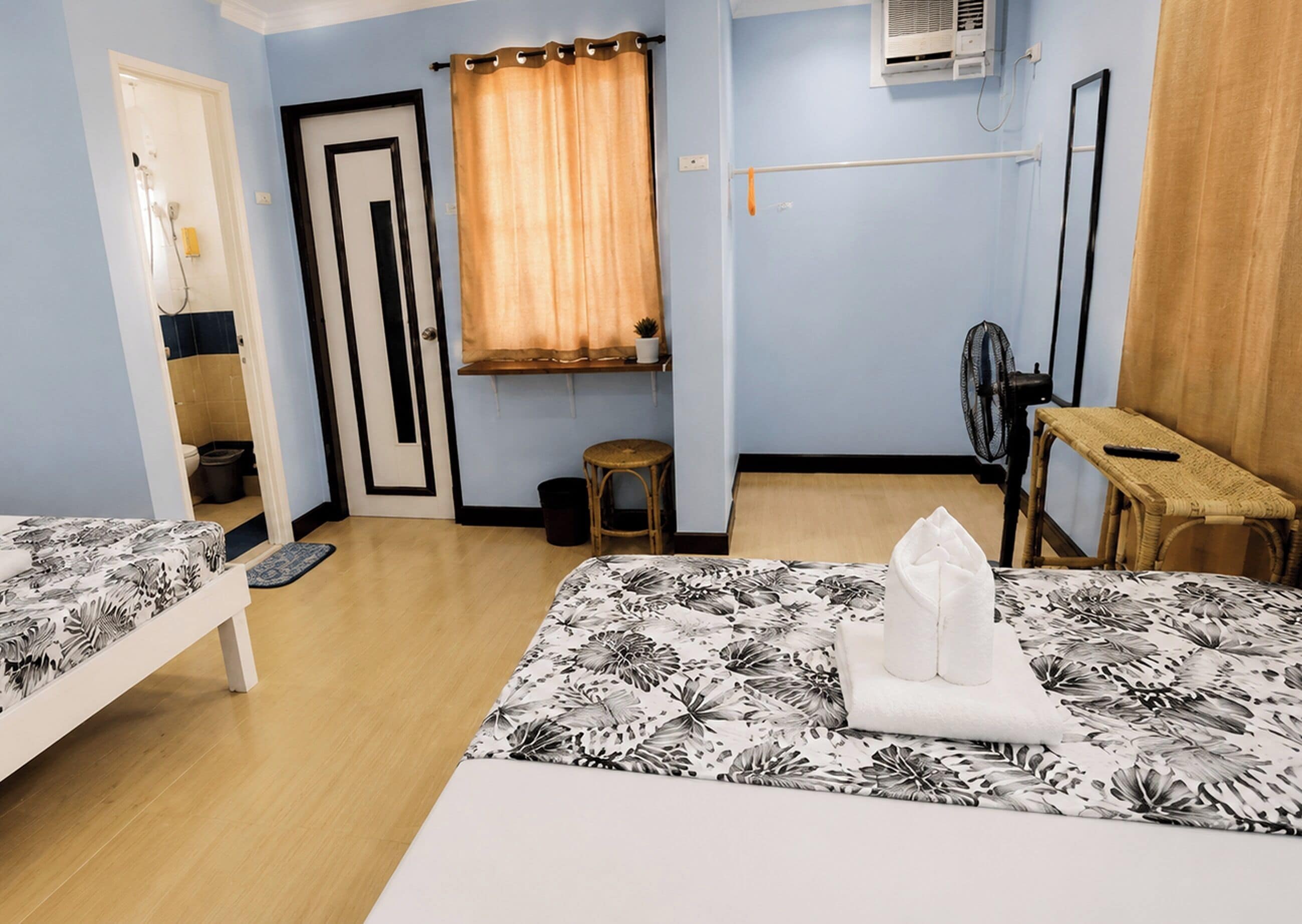 A room with two beds, a small stool, a chair, a fan, and a window with curtains.