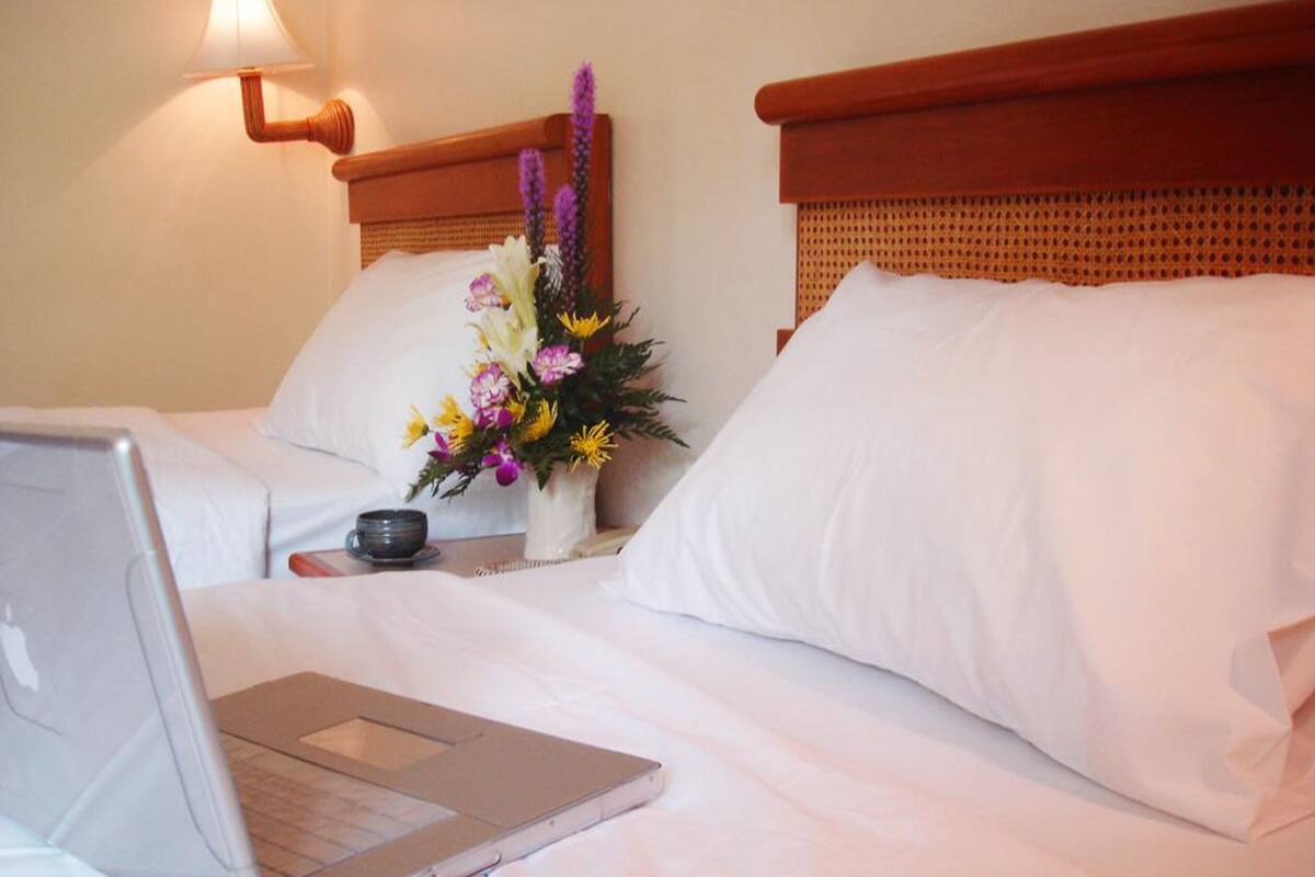 A hotel room with two beds, a laptop on one bed, a vase with flowers, and a lamp on the wall.