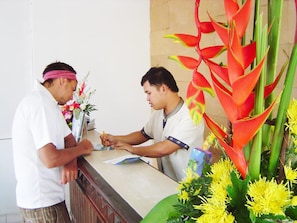 A reception desk with a man and a woman standing behind it, a plant with red and yellow leaves, and a yellow flower arrangement.