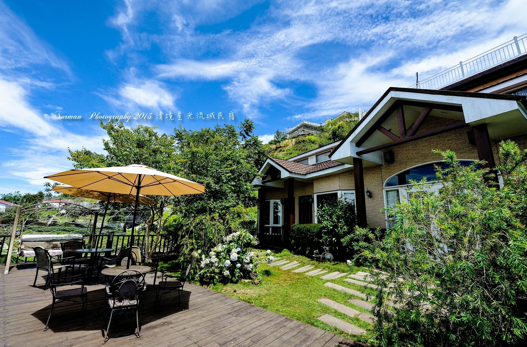 Un patio con mobiliario exterior y un gran paraguas, rodeado de follaje y una casa con techo inclinado.