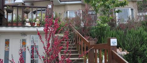En boligbygning med en trætrap, der fører op til en balkon, omgivet af forskellige planter og blomster.