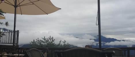 An outdoor patio with wicker chairs and large beige umbrellas, overlooking a mountainous landscape.