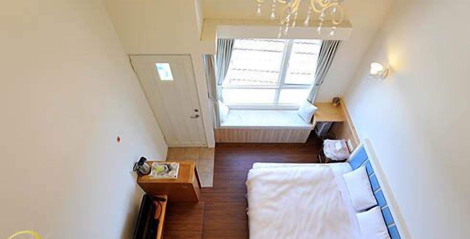 A small, compact room with a bed, a bedside table, a window with curtains, and a door.