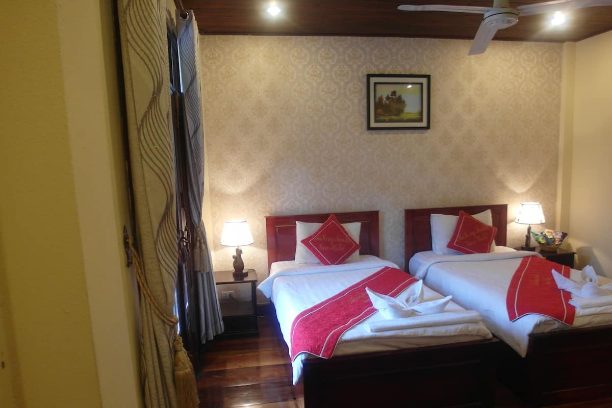 A hotel room with two beds, wooden floors, a ceiling fan, a framed picture on the wall, and a nightstand with a lamp and snacks.