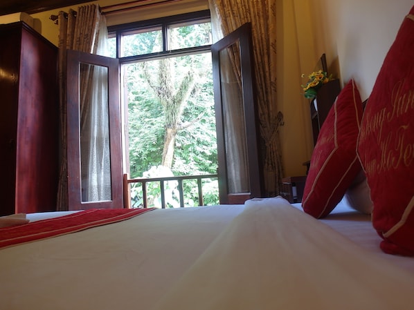 A room with a large bed, a view of trees through an open window, and a red decorative pillow.