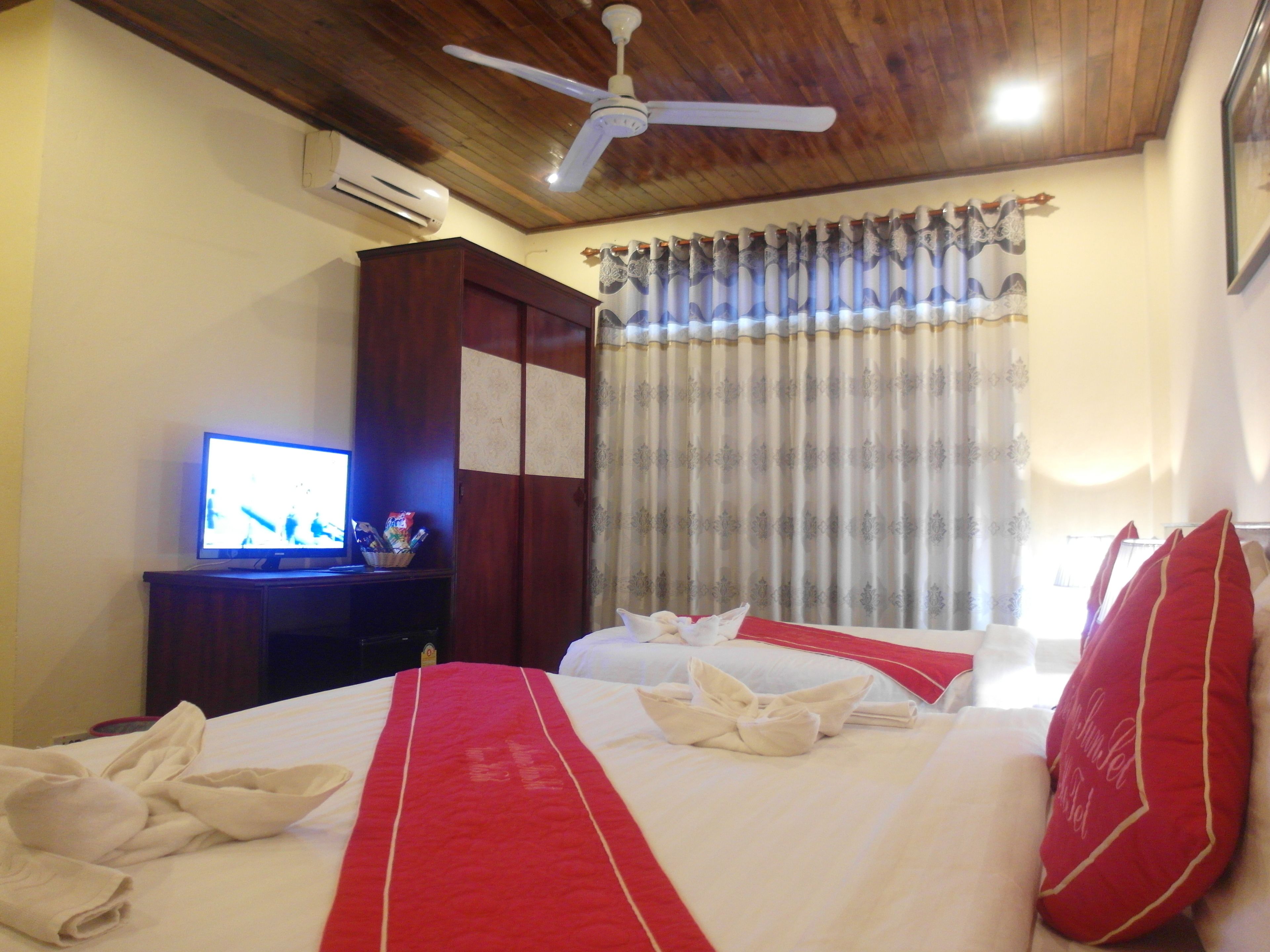 A hotel room with two beds, a television, an air conditioning unit, a wardrobe, and a ceiling fan.
