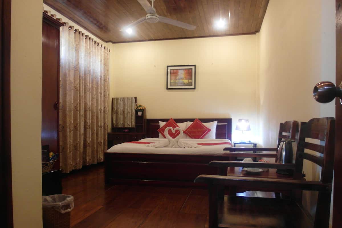 A hotel room with a wooden floor, a bed with white bedding and red pillows, a desk with a lamp, and a ceiling fan.
