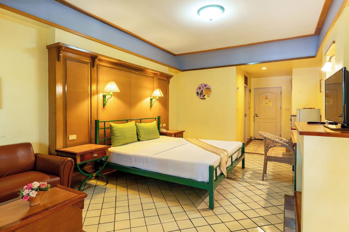A hotel room with a green bed, two bedside lamps, a wooden headboard, a green chair, a small table with a vase of flowers, and a tiled floor.