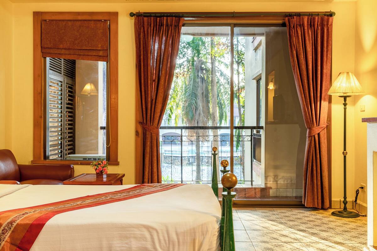 A hotel room with a bed, a window with curtains, a lamp, and a view of the outside.