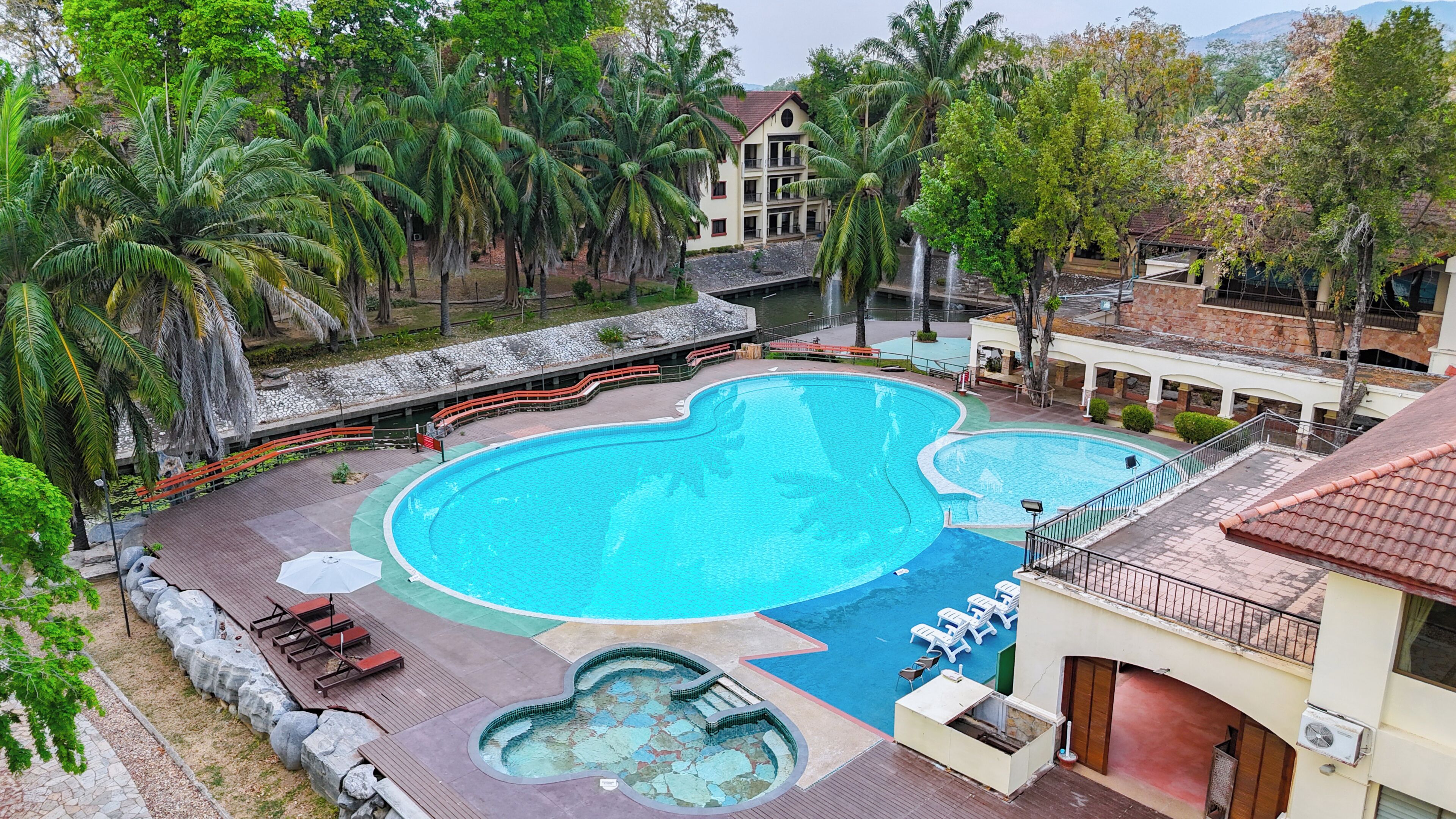 A large swimming pool with a unique shape, surrounded by palm trees and a building with multiple balconies.