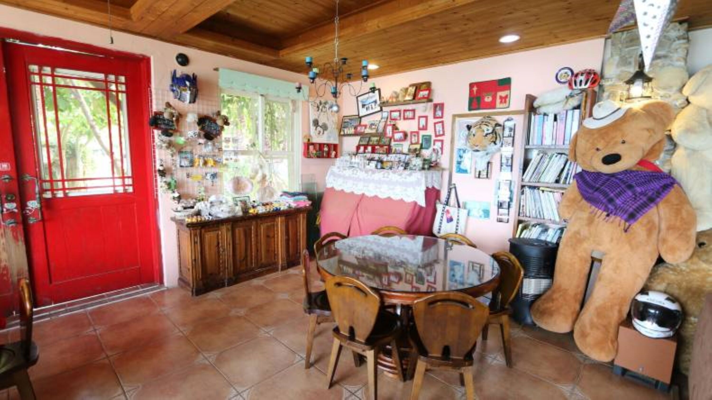 A cozy dining area with a red door, a large teddy bear, and a bookshelf filled with books and decorations.