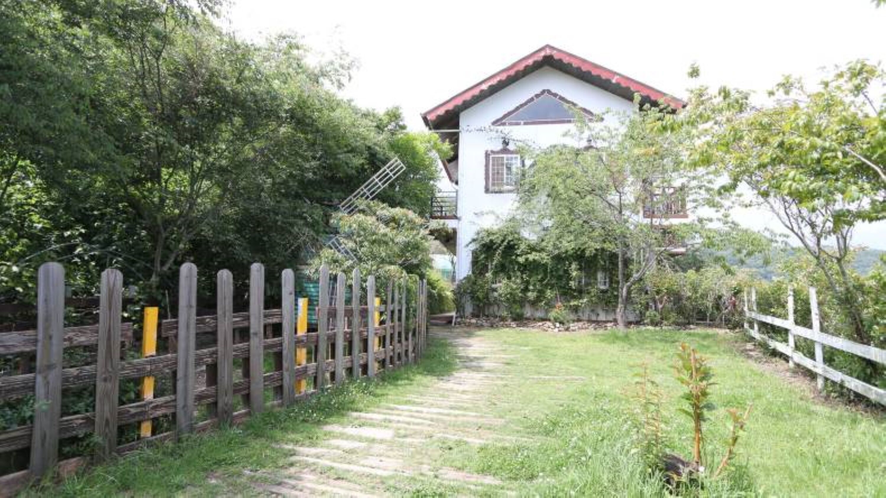 A white building with a red roof, surrounded by a wooden fence and greenery.
