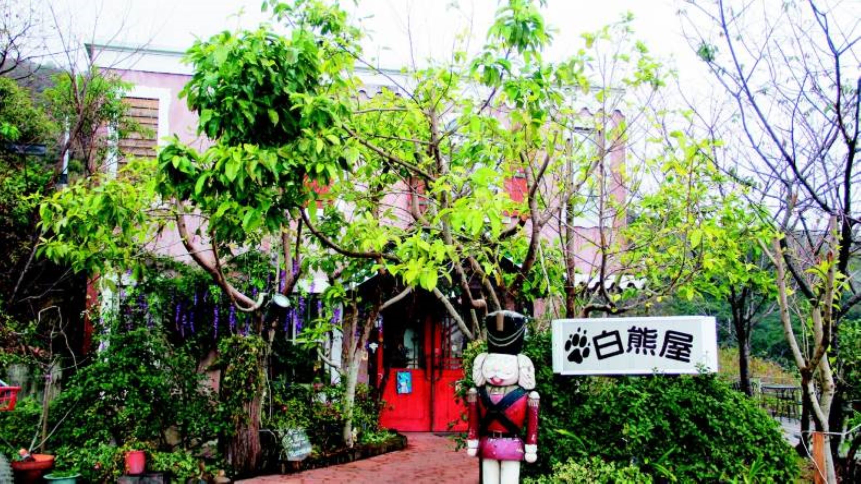 A building with a red door, surrounded by lush greenery and a sign with Japanese characters.