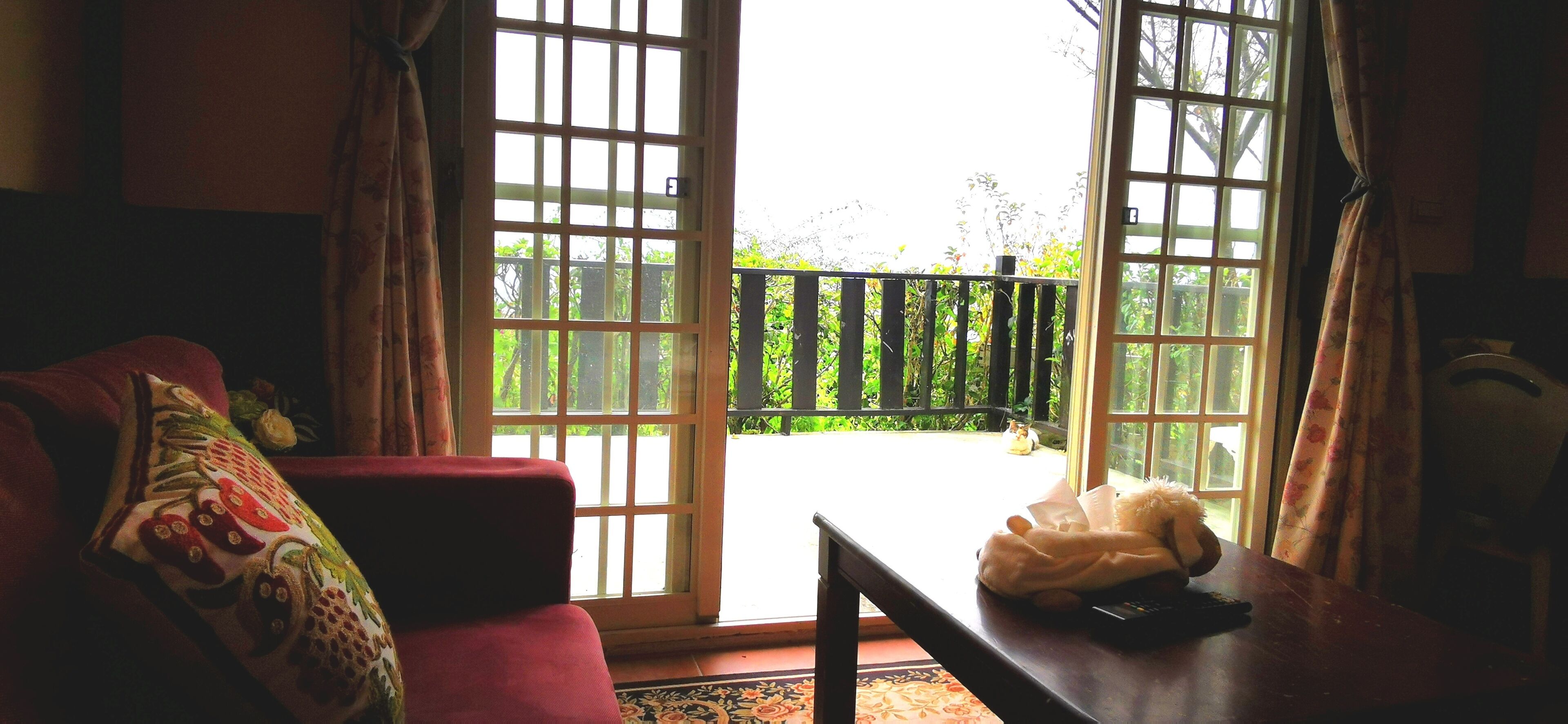 A room with a red sofa, a table with a remote control, and a view of a balcony with a wooden railing.