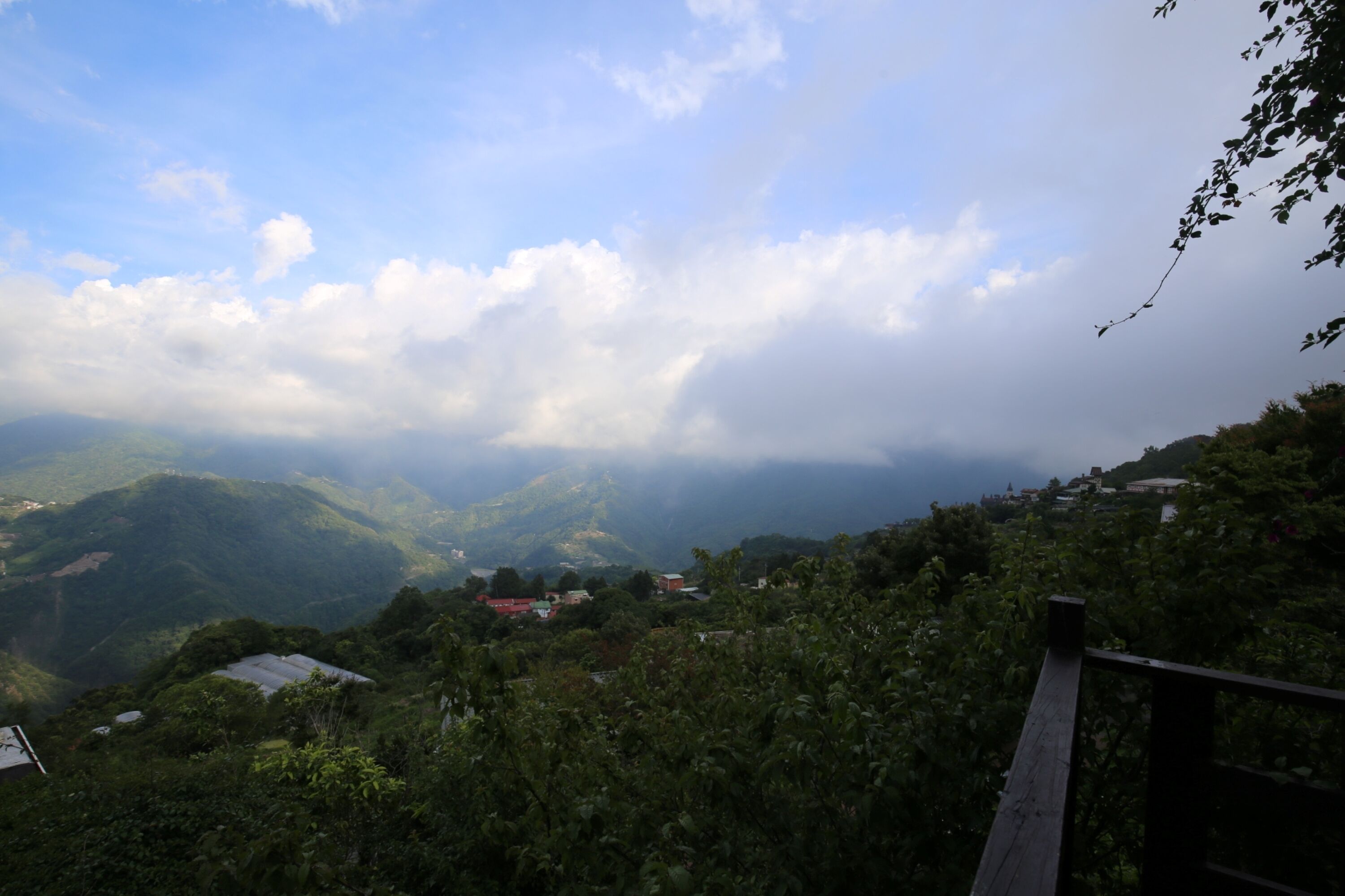 A mountainous landscape with a wooden railing, lush greenery, and a few buildings in the distance.