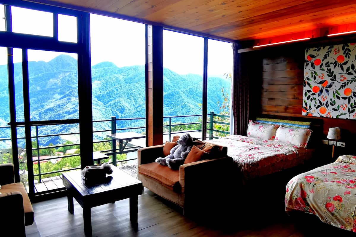 A room with a large window offering a mountain view, a sofa, a bed with floral bedding, and a wooden ceiling.