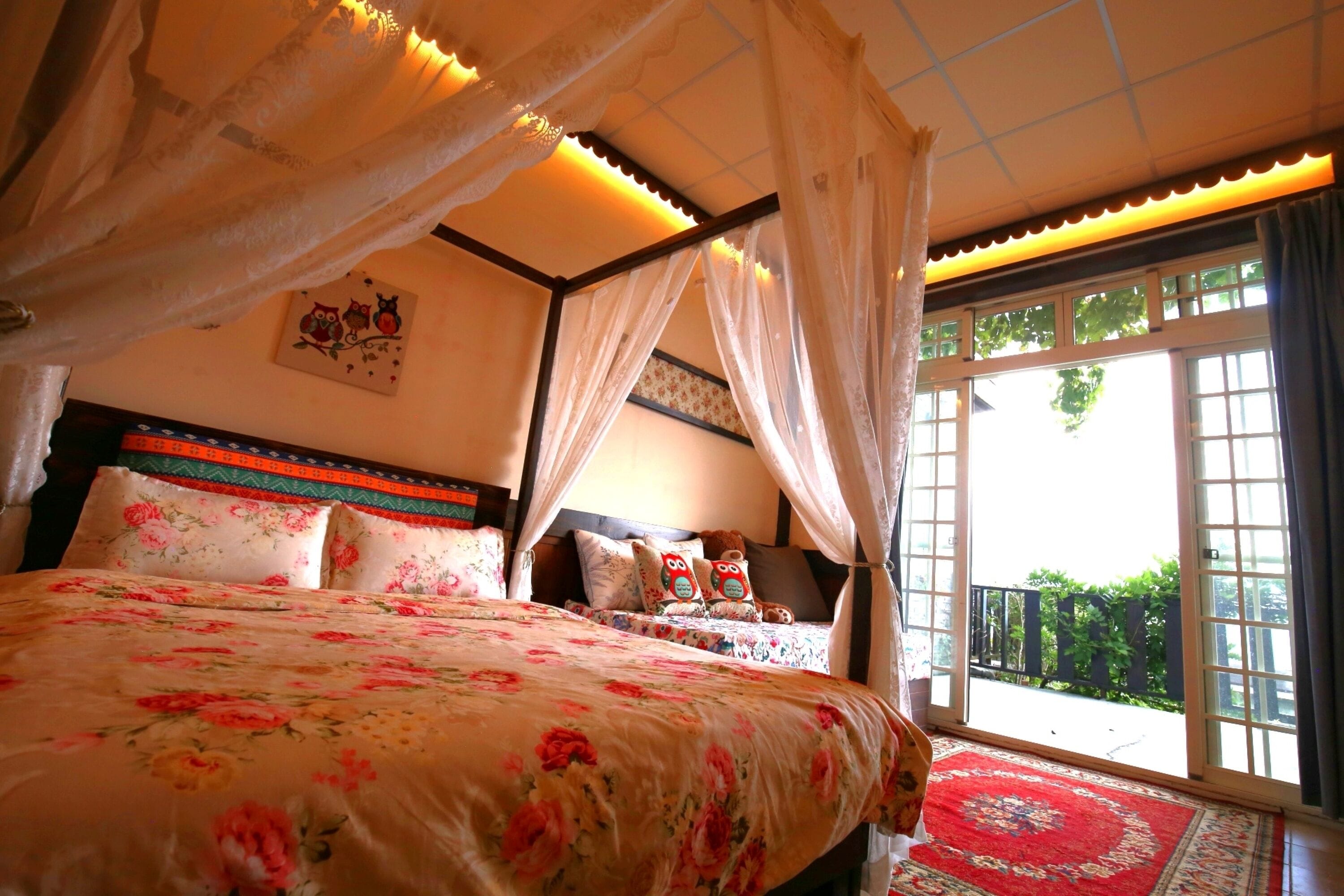 A bedroom with a large bed, floral bedding, a headboard with decorative pillows, a wall with a painting, and a balcony with a red rug.