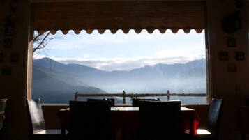 A dining area with a table and chairs, a wooden ceiling, and a view of mountains through a window with a scalloped edge.