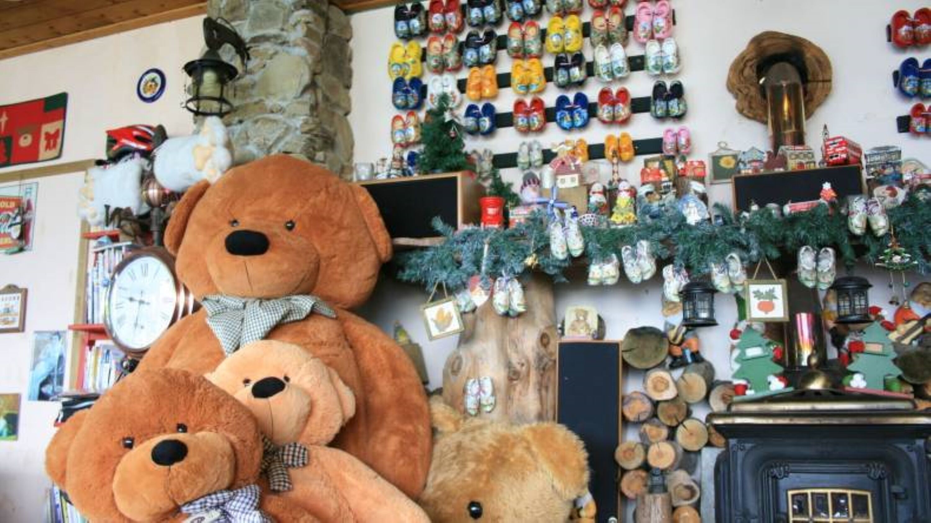 A room with a large teddy bear, a clock, and a wall decorated with various items including a Christmas tree and a collection of shoes.