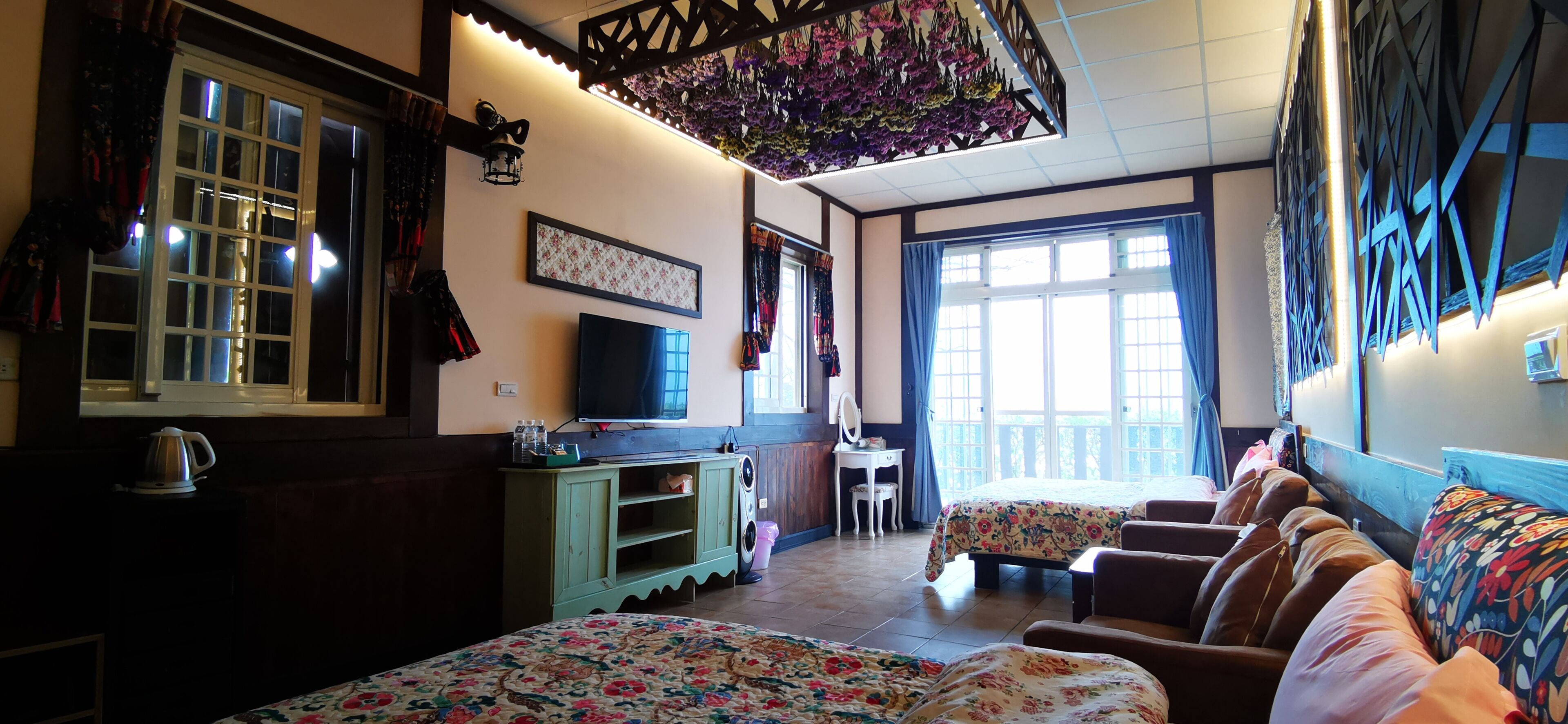 A cozy room with a bed, a desk, and a chair. There is a window with curtains, a wall-mounted TV, and a decorative hanging.