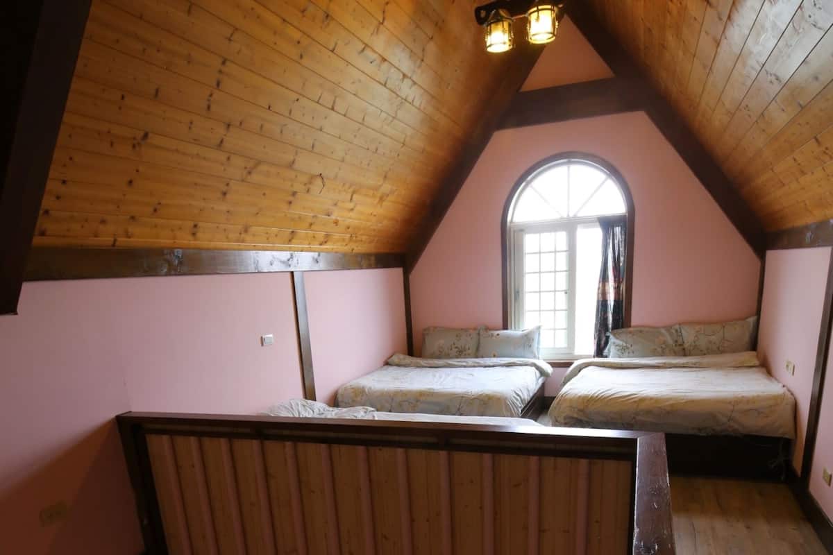 A cozy attic bedroom with wooden beams, two beds, a small table, and a window with curtains.