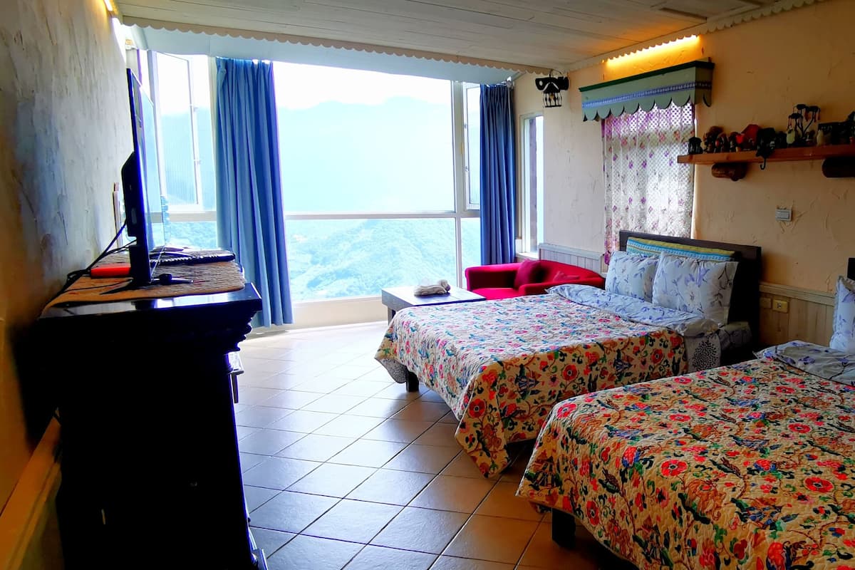 A room with two beds, a desk, and a chair. There is a window with curtains, a shelf with decorative items, and a view of the mountains.