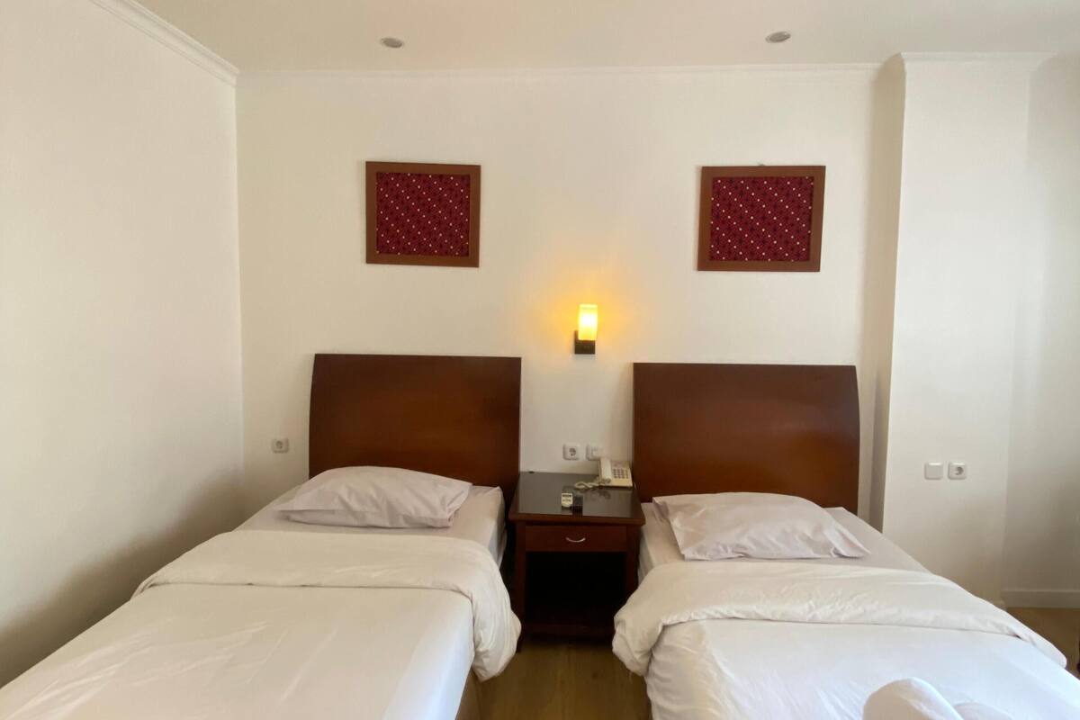Habitación de hotel con dos camas individuales, cabeceros de madera, mesitas de noche pequeñas con lámparas y dos marcos de cuadros colgados en la pared.