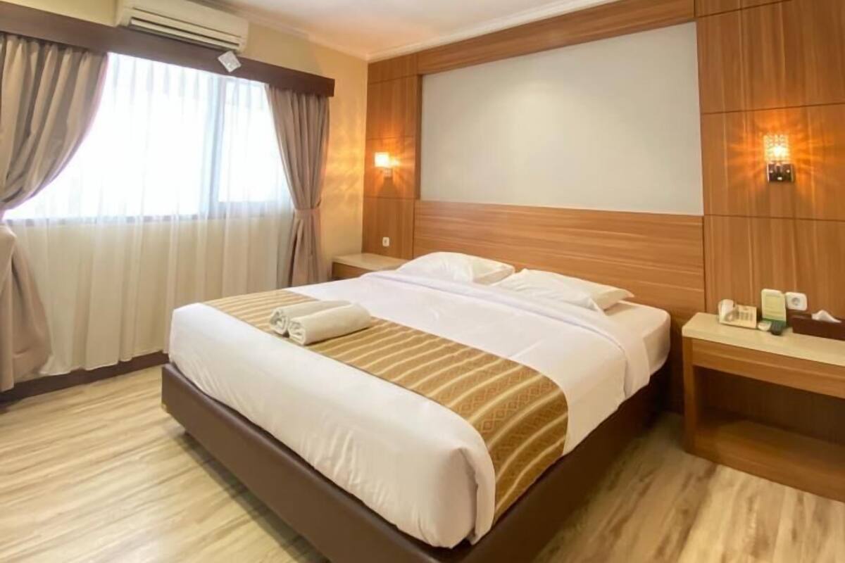 Una habitación de hotel con una cama grande, cabecera de madera, mesitas de noche y un equipo de aire acondicionado.