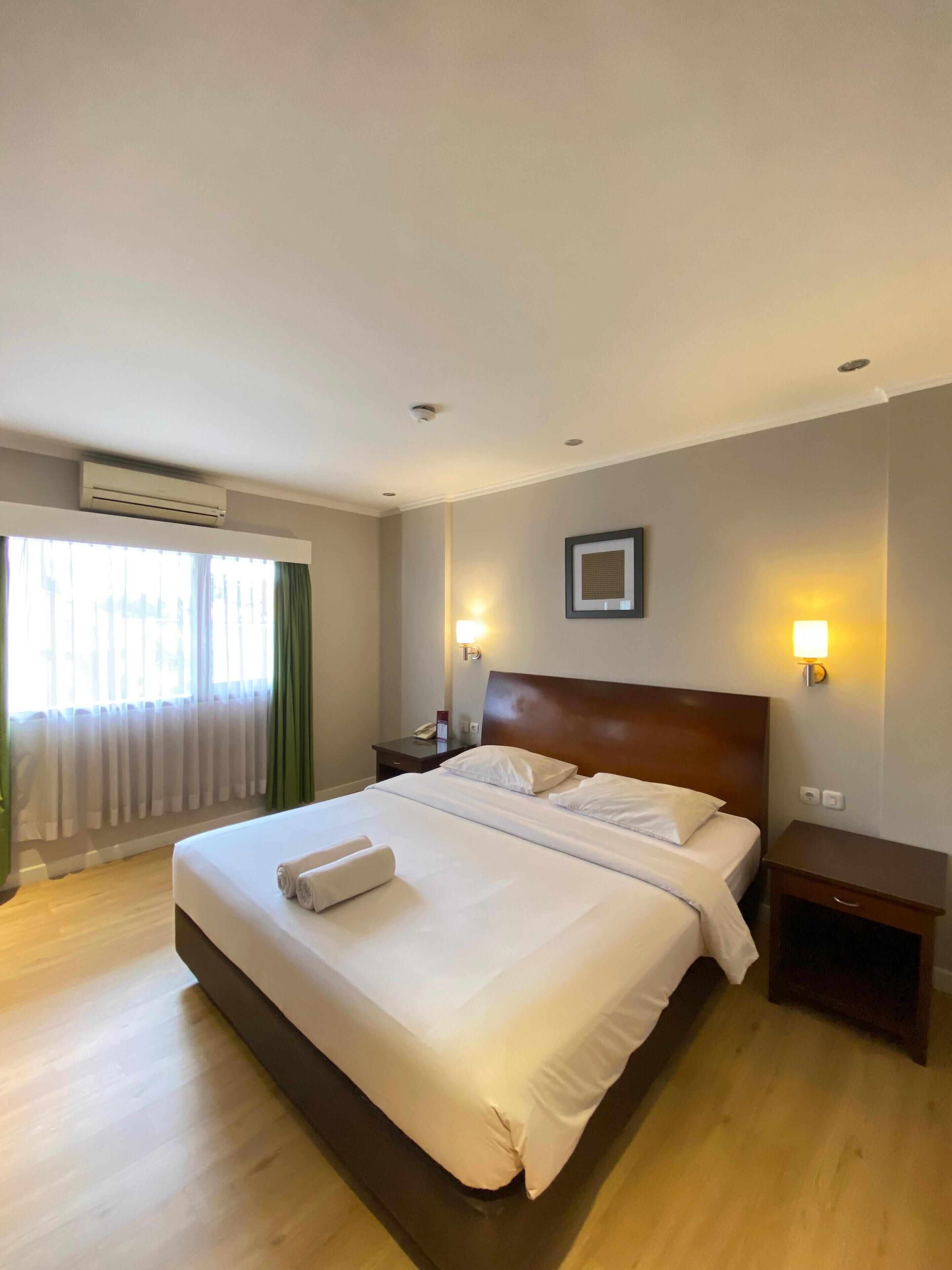 A hotel room with a large bed, two bedside tables, an air conditioning unit, and a window with curtains.