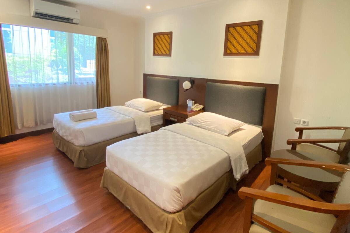 Habitación de hotel con dos camas, una silla de madera, un escritorio con teléfono y un equipo de aire acondicionado.