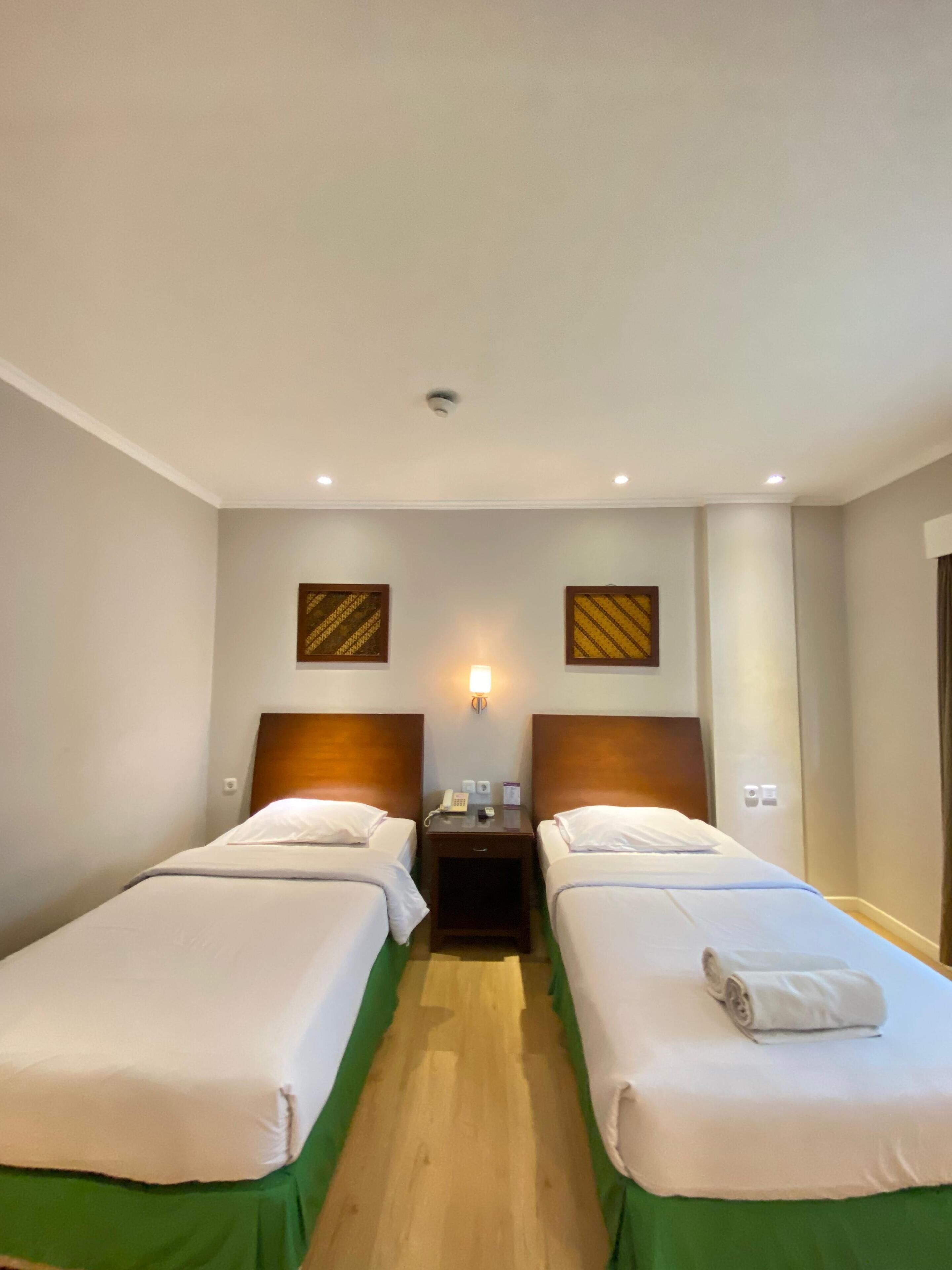 Habitación de hotel con dos camas individuales, una mesita de noche con teléfono y lámpara, y cabecera de madera.