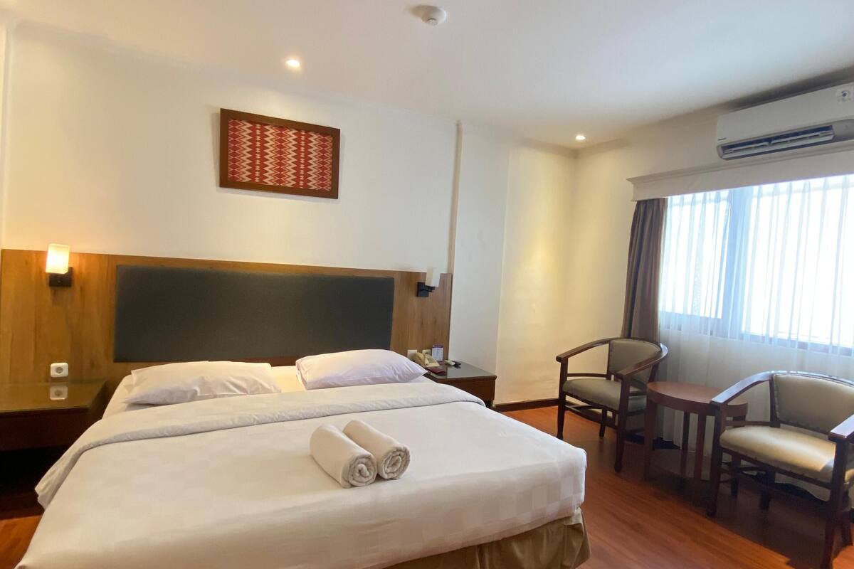 Habitación de hotel con una cama grande, un escritorio, dos sillas, aire acondicionado de pared y una ventana con cortinas.