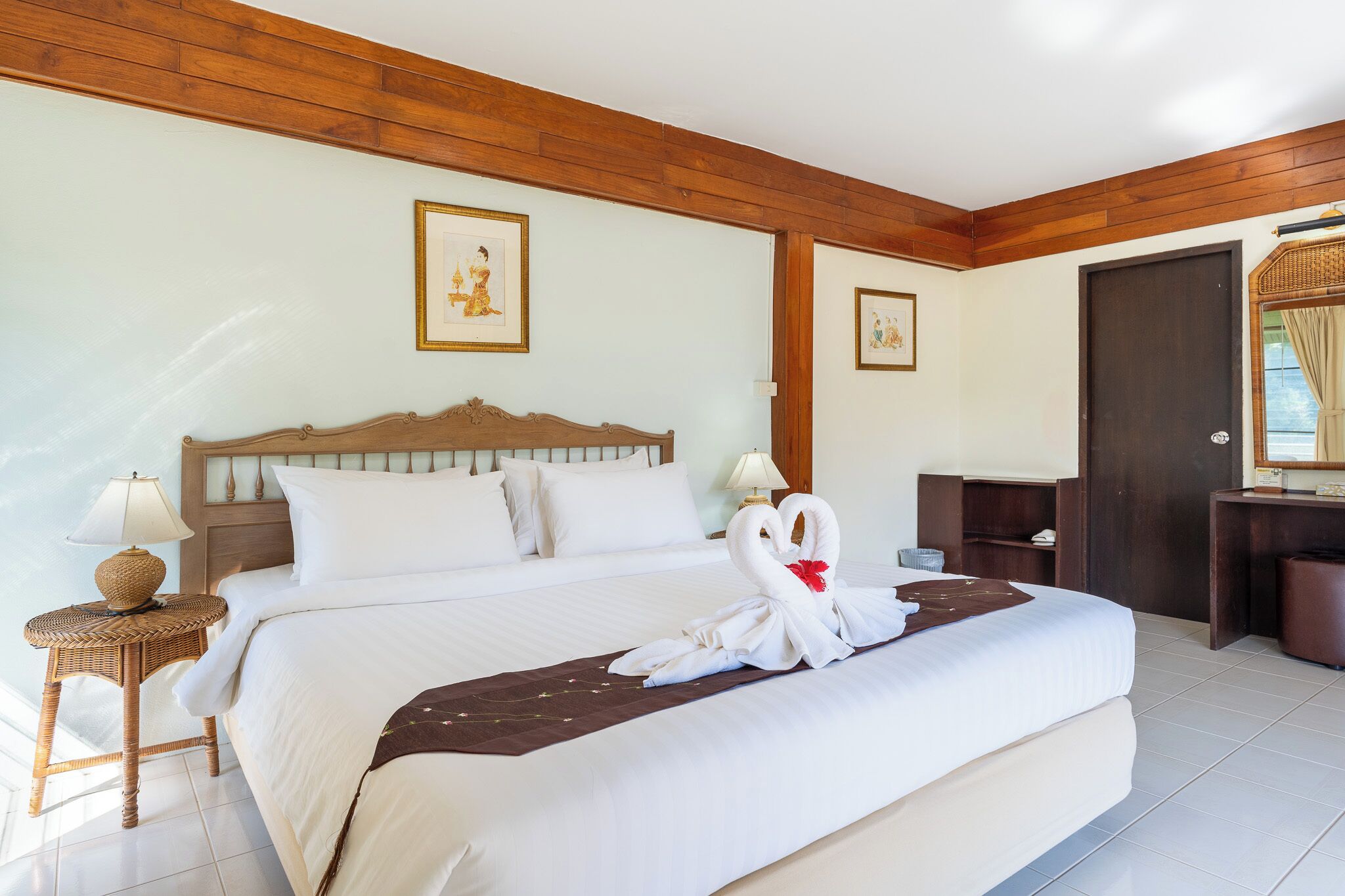 A hotel room with a large bed, wooden headboard, two framed pictures on the wall, a desk with a lamp, and a door with a view.