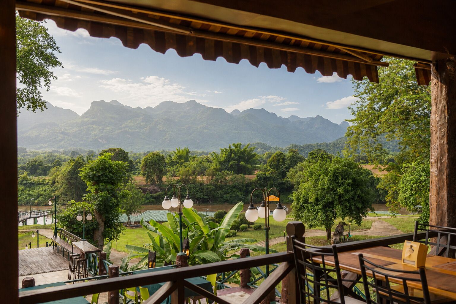 A terrace with a view of a river and mountains, wooden furniture, and a railing.