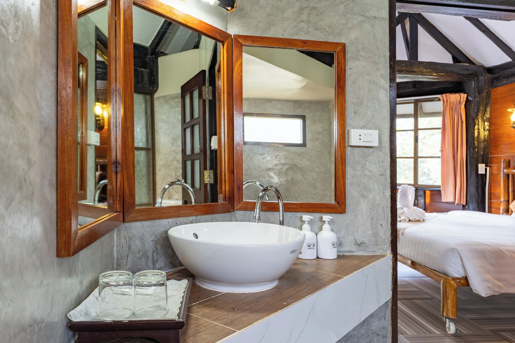 Un bagno con un lavandino ampio e bianco, un mobile contenitore in legno e uno specchio. Si intravede una porta aperta che dà su un letto con struttura in legno e biancheria da letto bianca.