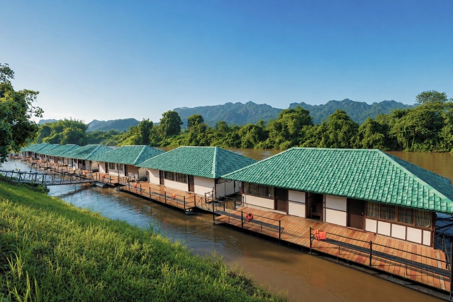 A row of traditional-style buildings with green roofs, wooden walkways, and a river in the foreground.