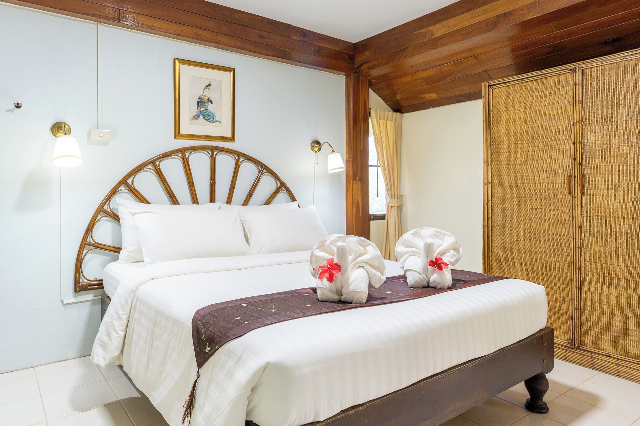A neatly made bed with white linens and two towels with red ribbons, a wooden headboard, a framed picture on the wall, and a large woven wardrobe.
