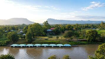 A riverside hotel with a row of rooms along the water, a park area, and distant mountains.