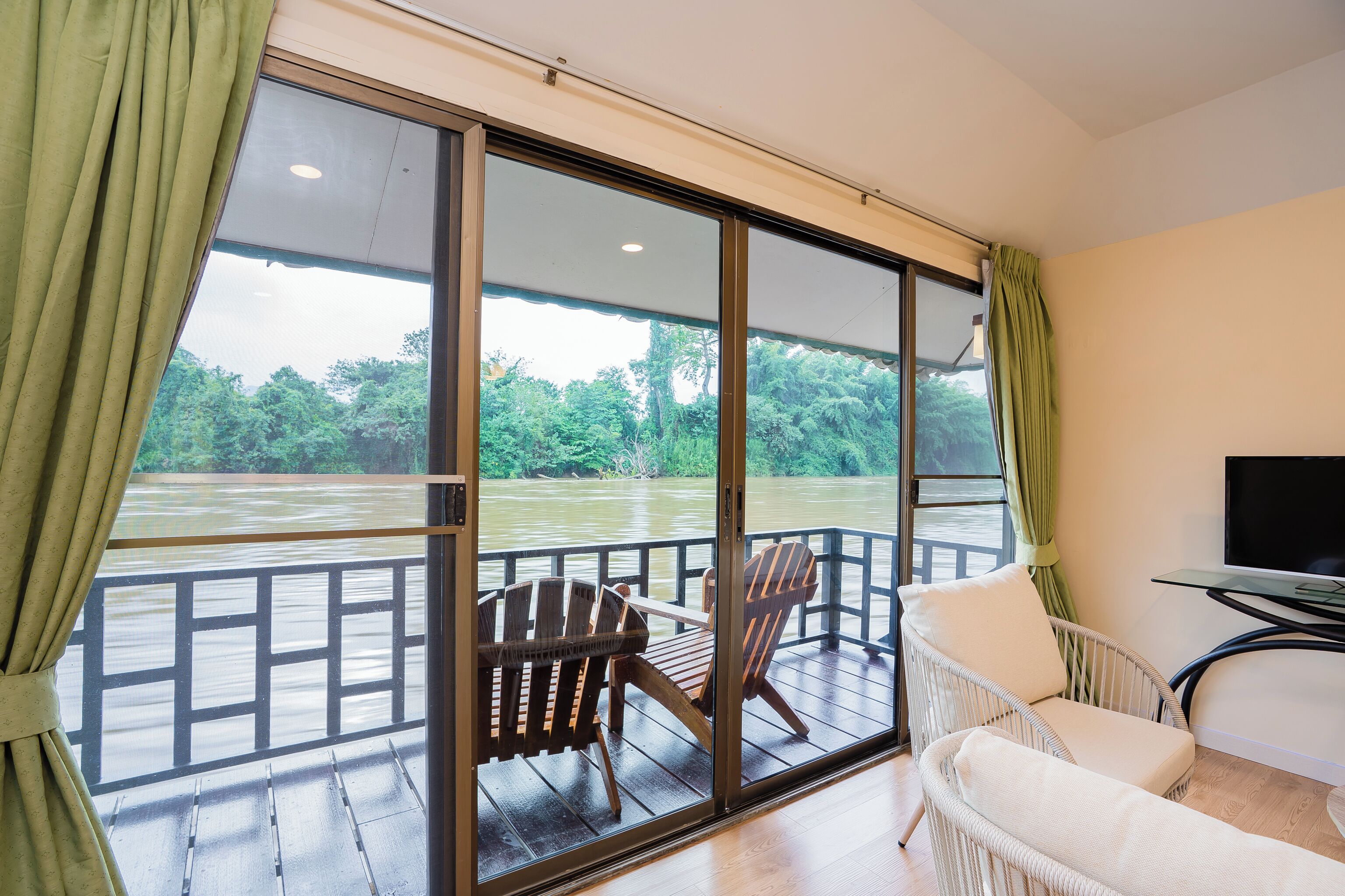 A room with a large window overlooking a river, a wooden deck, and a television on a stand.