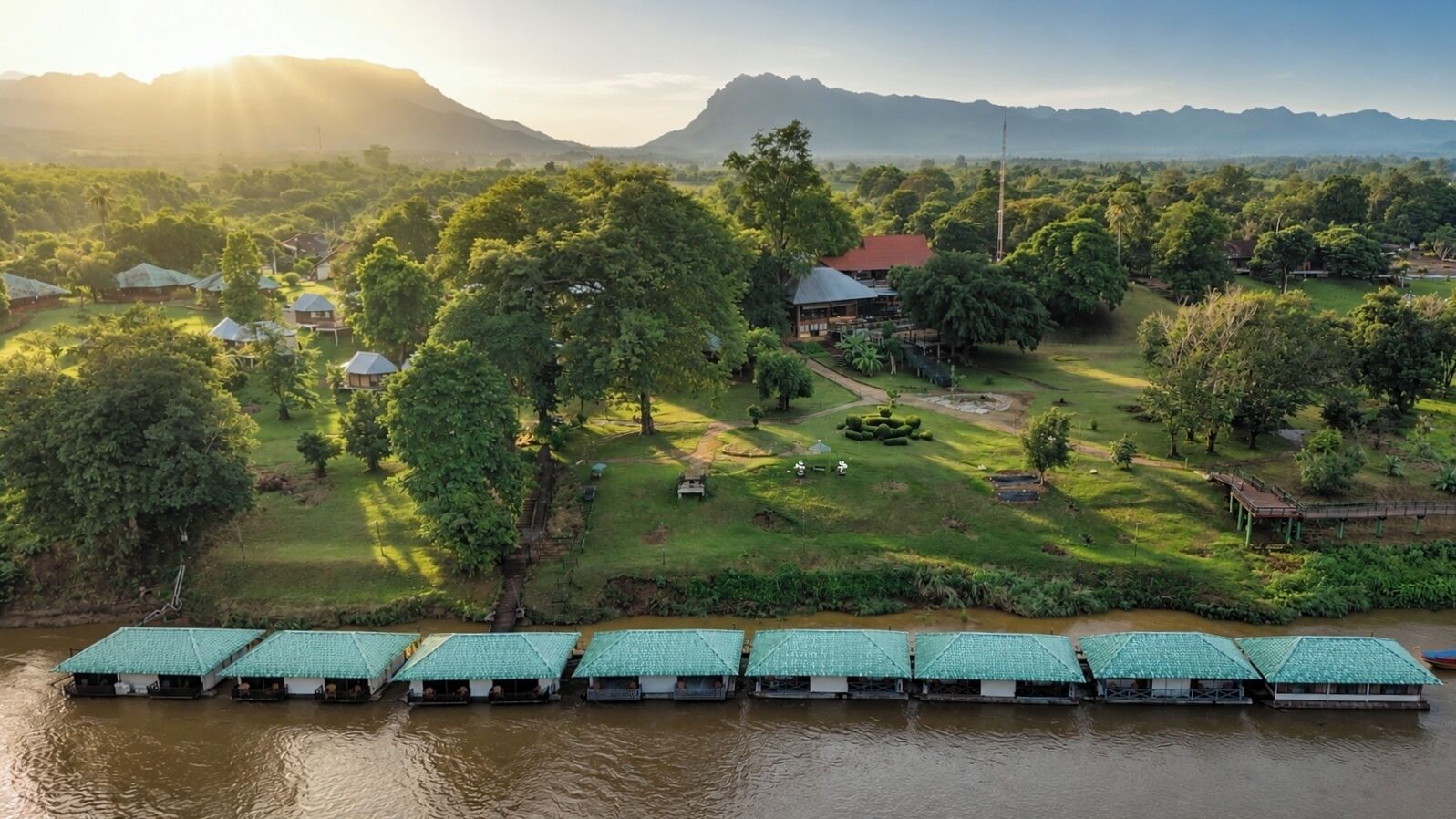 A riverside accommodation with floating structures, surrounded by lush greenery and distant mountains.