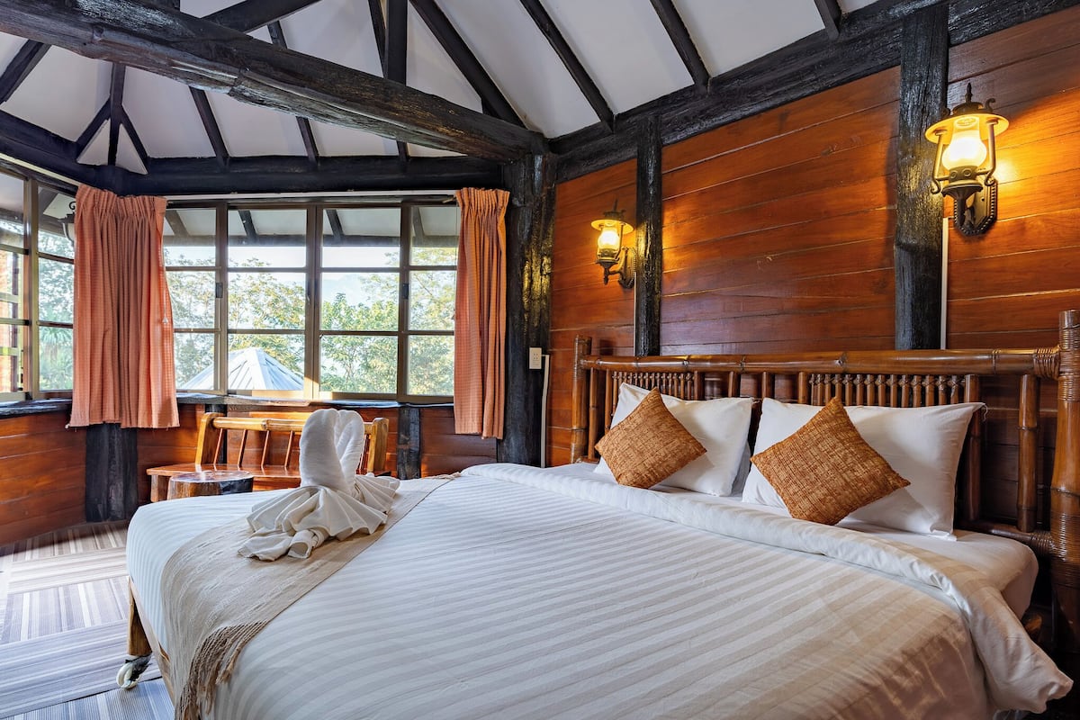 A large bed with white linens and two pillows, a wooden headboard, and a window with curtains.