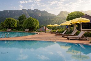Un área de piscina con sillas de descanso y sombrillas, rodeada de vegetación y montañas.