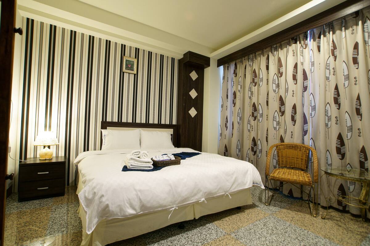 A hotel room with a large bed, a nightstand with a lamp, a wicker chair, and curtains with a leaf pattern.
