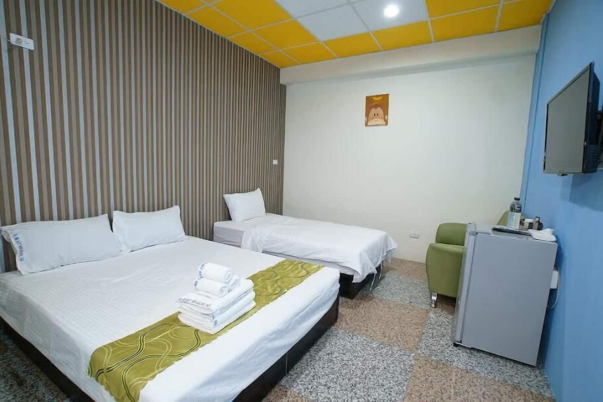 A hotel room with two beds, a green chair, a small desk with a computer, and a TV mounted on the wall.