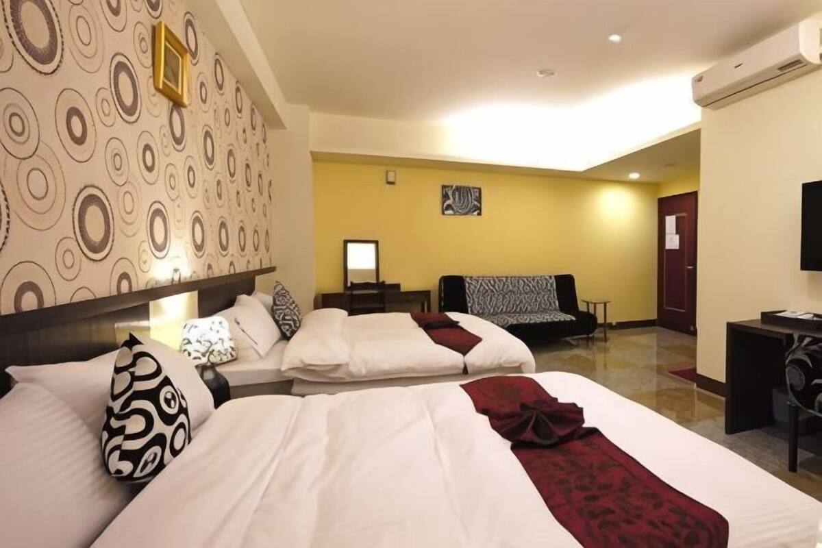 A hotel room with two beds, a desk, a chair, a TV, and a patterned wall.