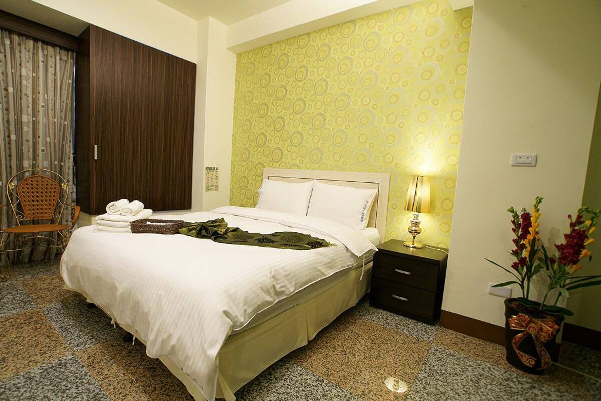A hotel room with a bed, a nightstand with a lamp, a chair, a potted plant, and a curtain.