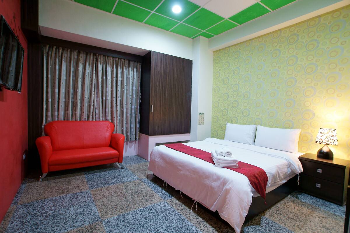 A hotel room with a bed, a red sofa, a nightstand with a lamp, and a patterned wall.